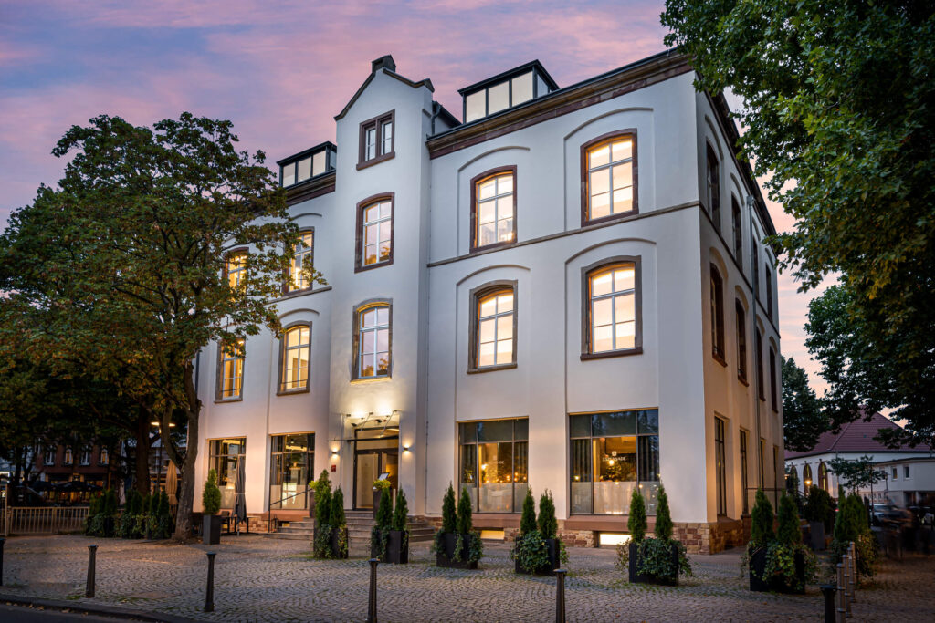 Hotel ESPLANADE – Aussenansicht. Es zeigt die Frontansicht des ESPLANADE-Hotels von draussen. Gruene Baeume umranden das Hotel und im im Hintergrund sieht man den Himmel im Dämmerlicht. Die Datei ist ein Foto im JPEG-Format.